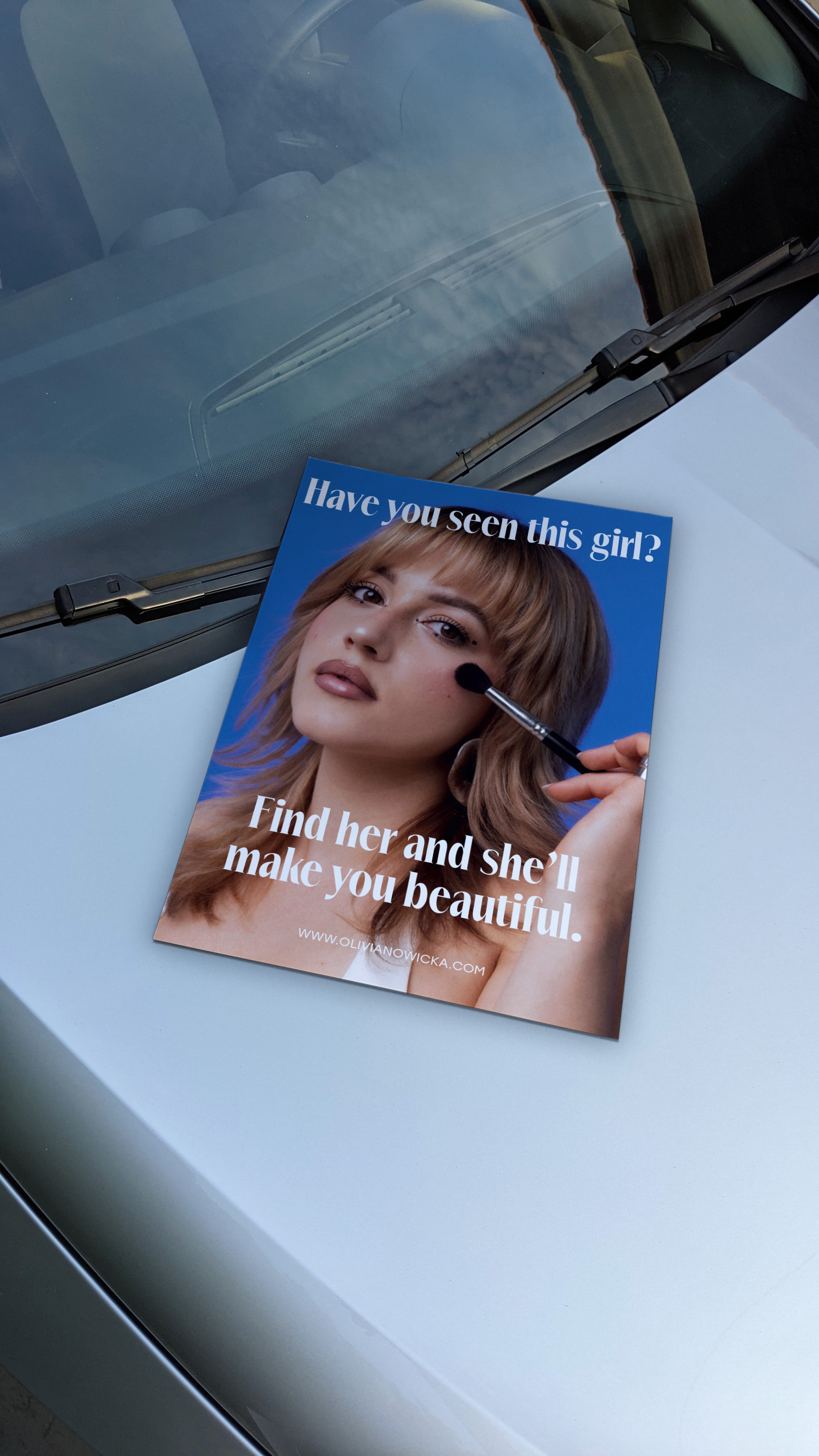 Makeup artist’s poster on a car hood, featuring a woman with glam makeup and text promoting her services with a website link.