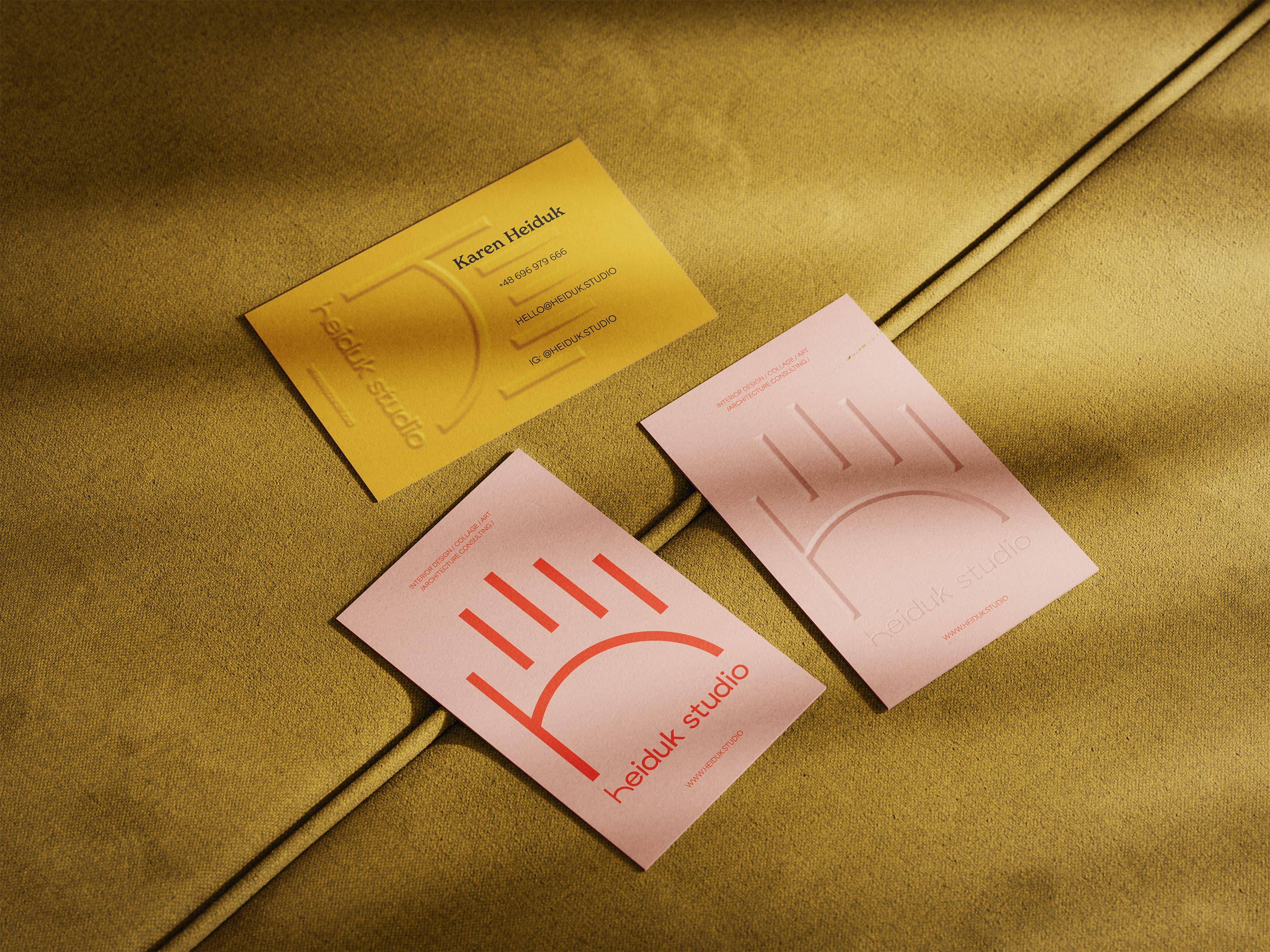 Three business cards for Heiduk Studio, two pink with a red logo and one mustard yellow, placed on textured fabric.